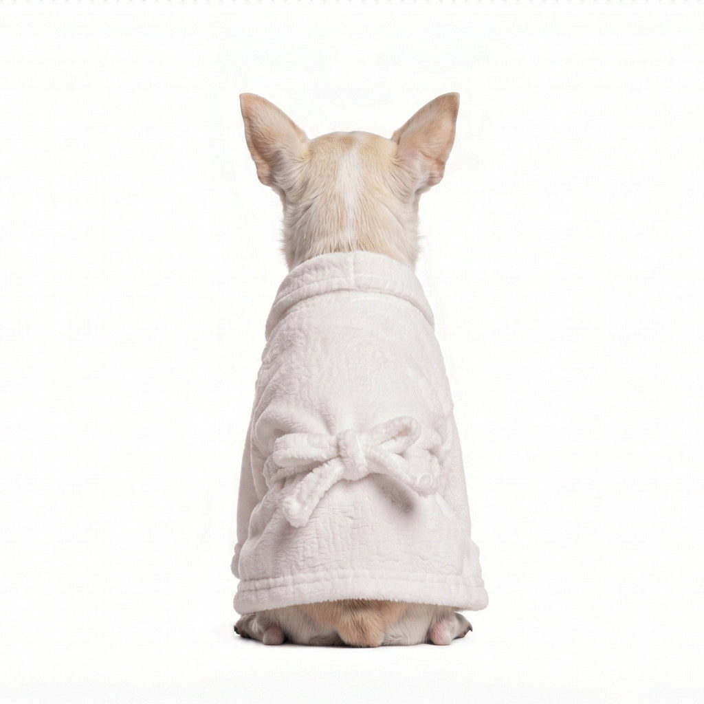 A small beige dog seen from behind, wearing a soft white plush bathrobe with a bow detail on the back, isolated on a white background.