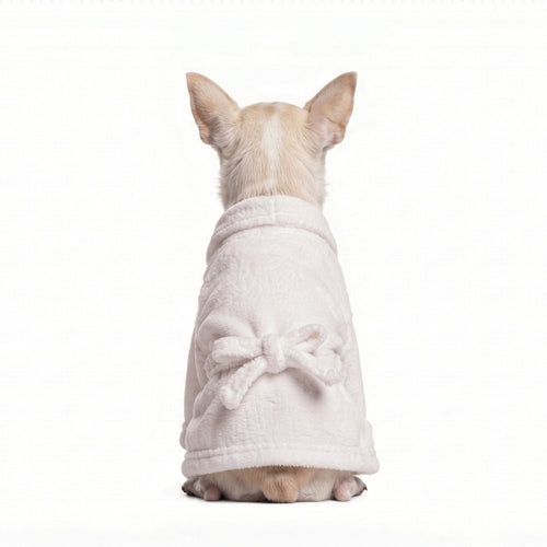 A small beige dog seen from behind, wearing a soft white plush bathrobe with a bow detail on the back, isolated on a white background.