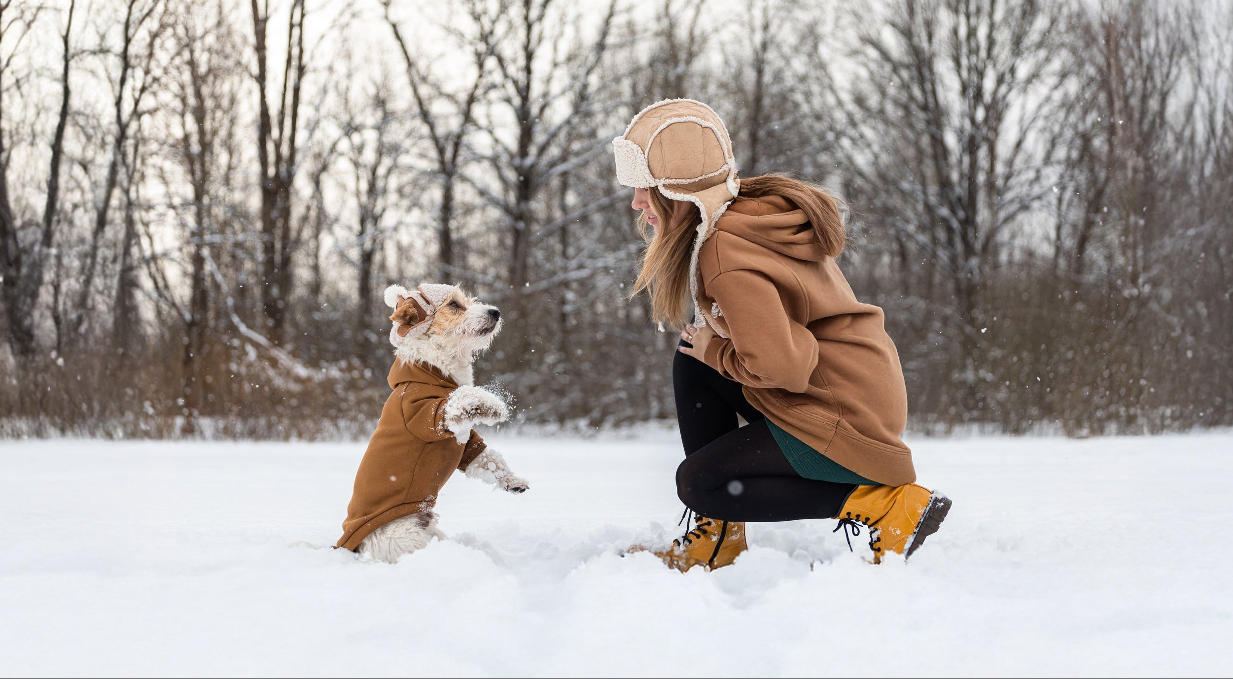 A woman in a brown winter coat and beige hat crouches in the snow while playing with a small dog wearing a matching brown coat in an outdoor winter setting.