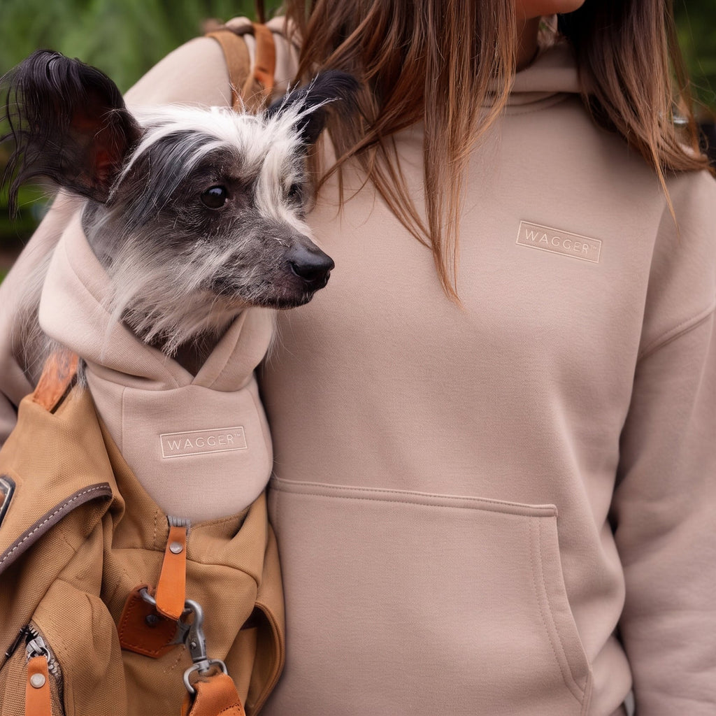 A small black and white dog wearing a tan backpack with a matching beige hooded insert, carried on the shoulders of a woman in a beige hoodie in an outdoor park setting.